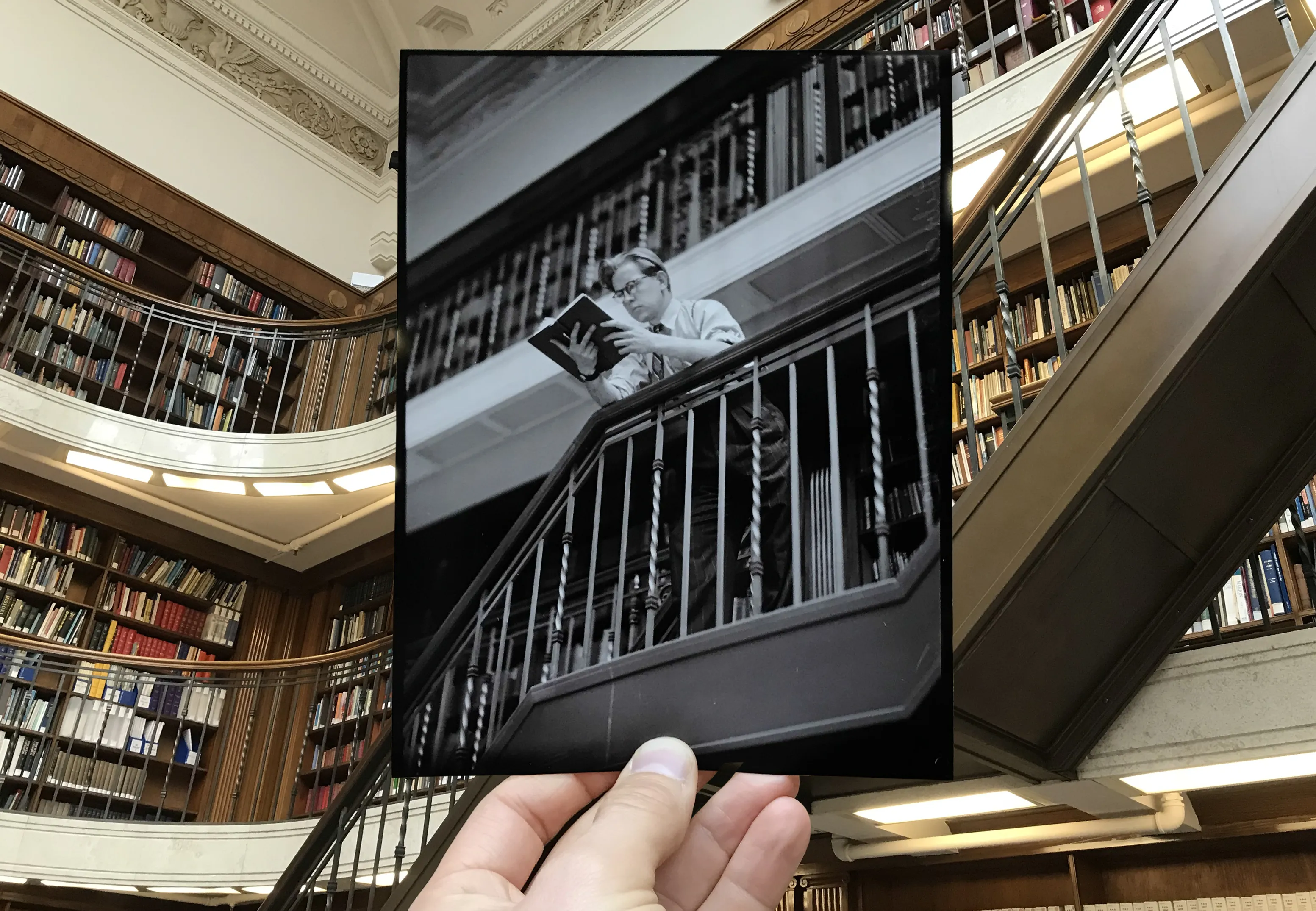 An old photograph of a man reading a book in the reading room, held up against the same background location where it was photographed decades earlier.