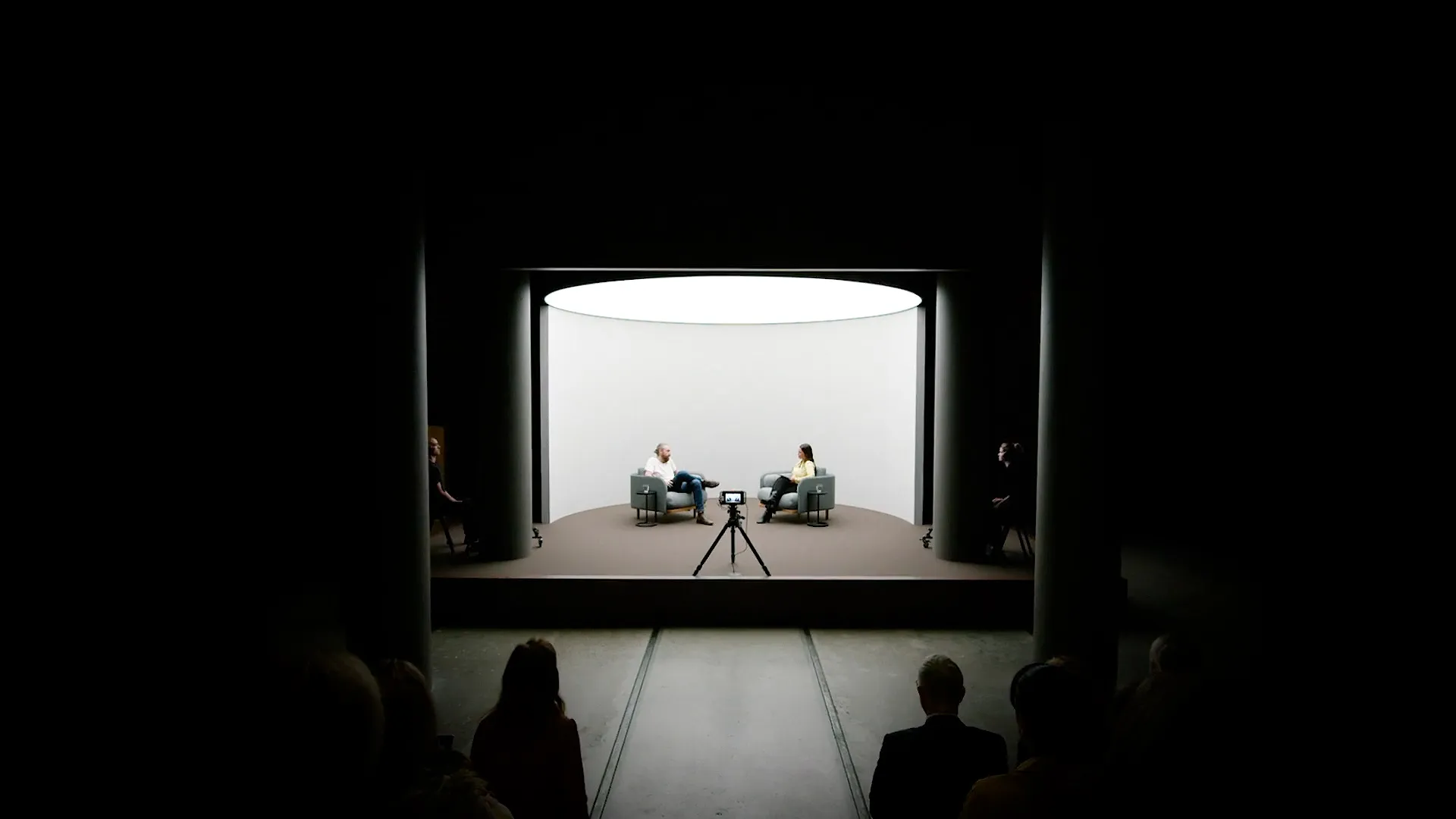 Two people seated opposite each other in a brightly lit circular interview space, viewed from the audience.