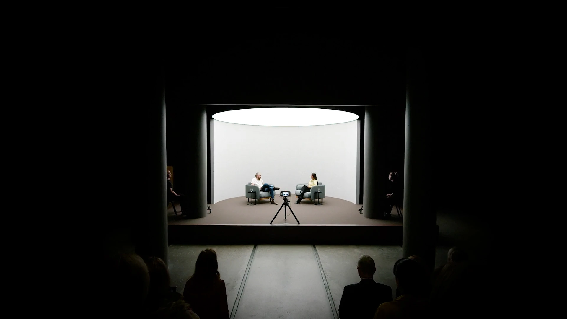 Two people seated opposite each other in a brightly lit circular interview space, viewed from the audience.