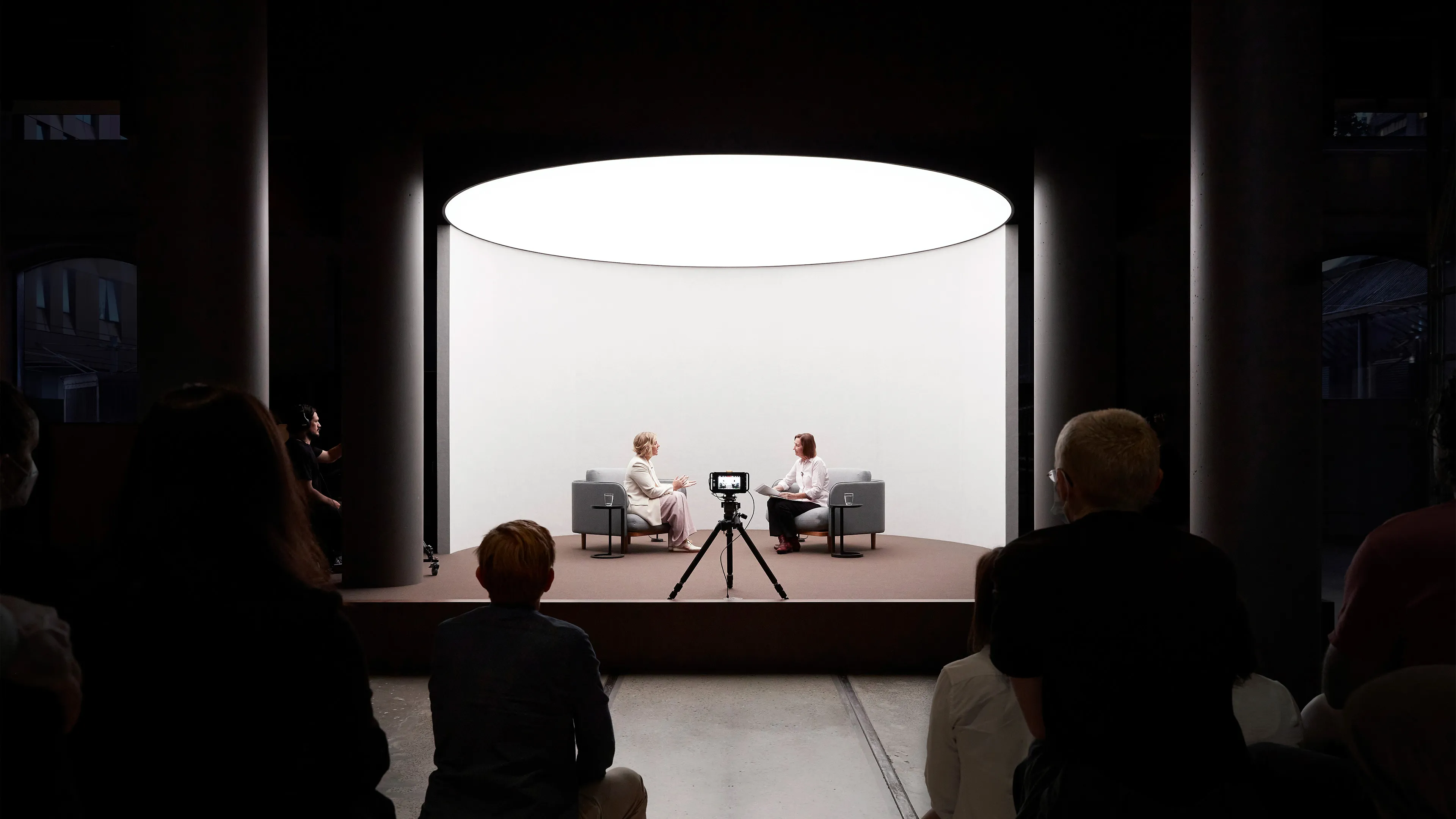 Two women sitting in large grey chairs on a well-lit stage conducting an interview. A camera on the stage films them while an audience watches in the foreground of the photo.