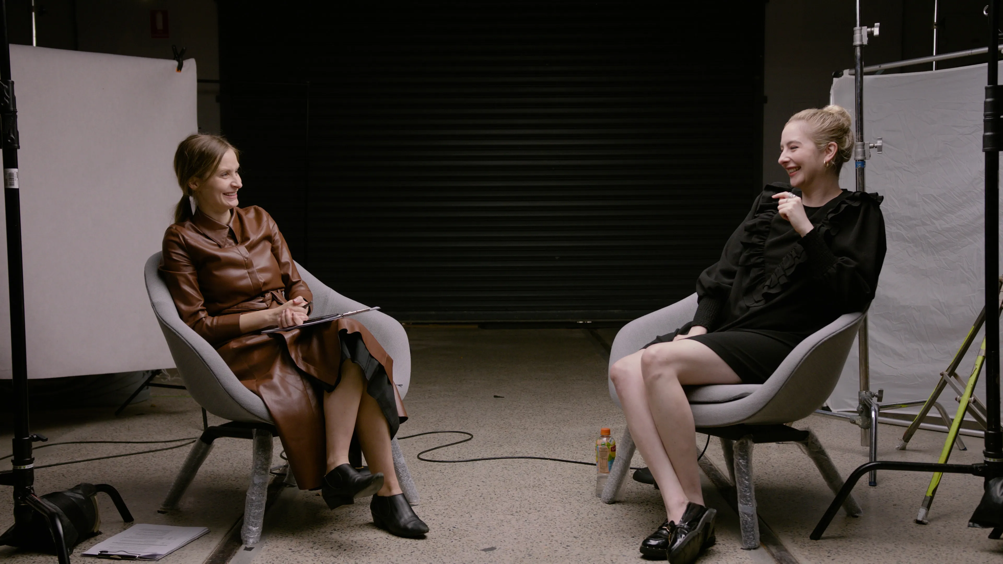 Two women, smiling at each other, sit on chairs, surrounded by lighting equipment.