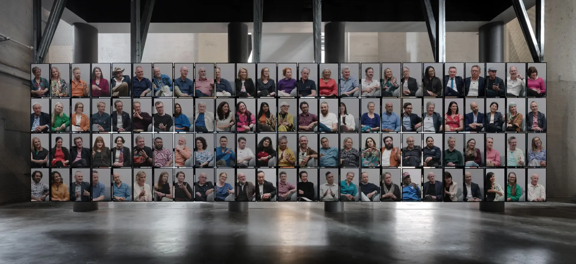 A grid of 100 screens hang from the ceiling in a large empty space. The grid is 25 screens wide and 4 screens tall.