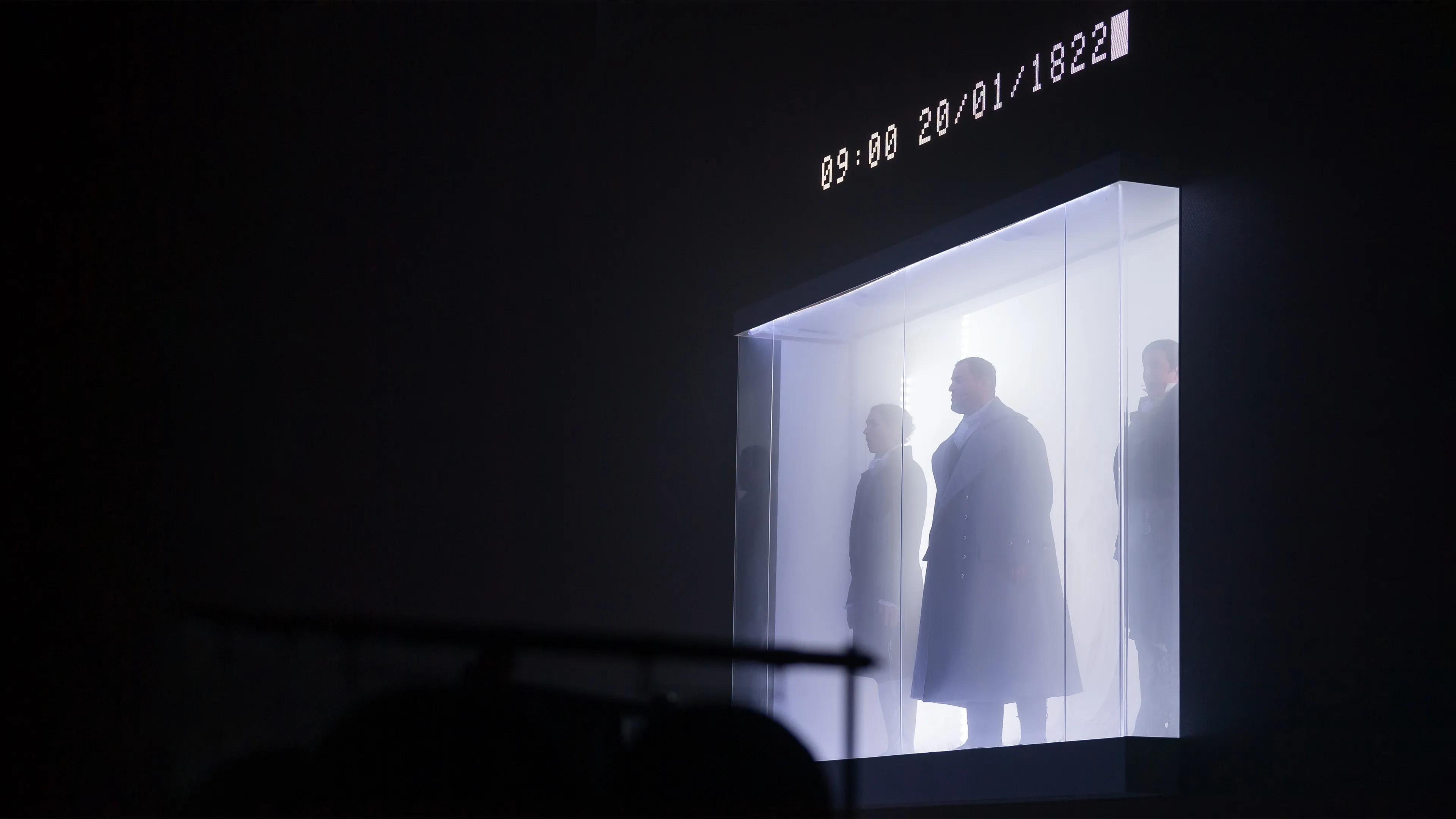 The time and date 09:00 20/01/1822 is displayed in large text on an LED screen that is surrounding a glass encloure. The enclosure is filled with fog and contains three opera performers singing in period costume.