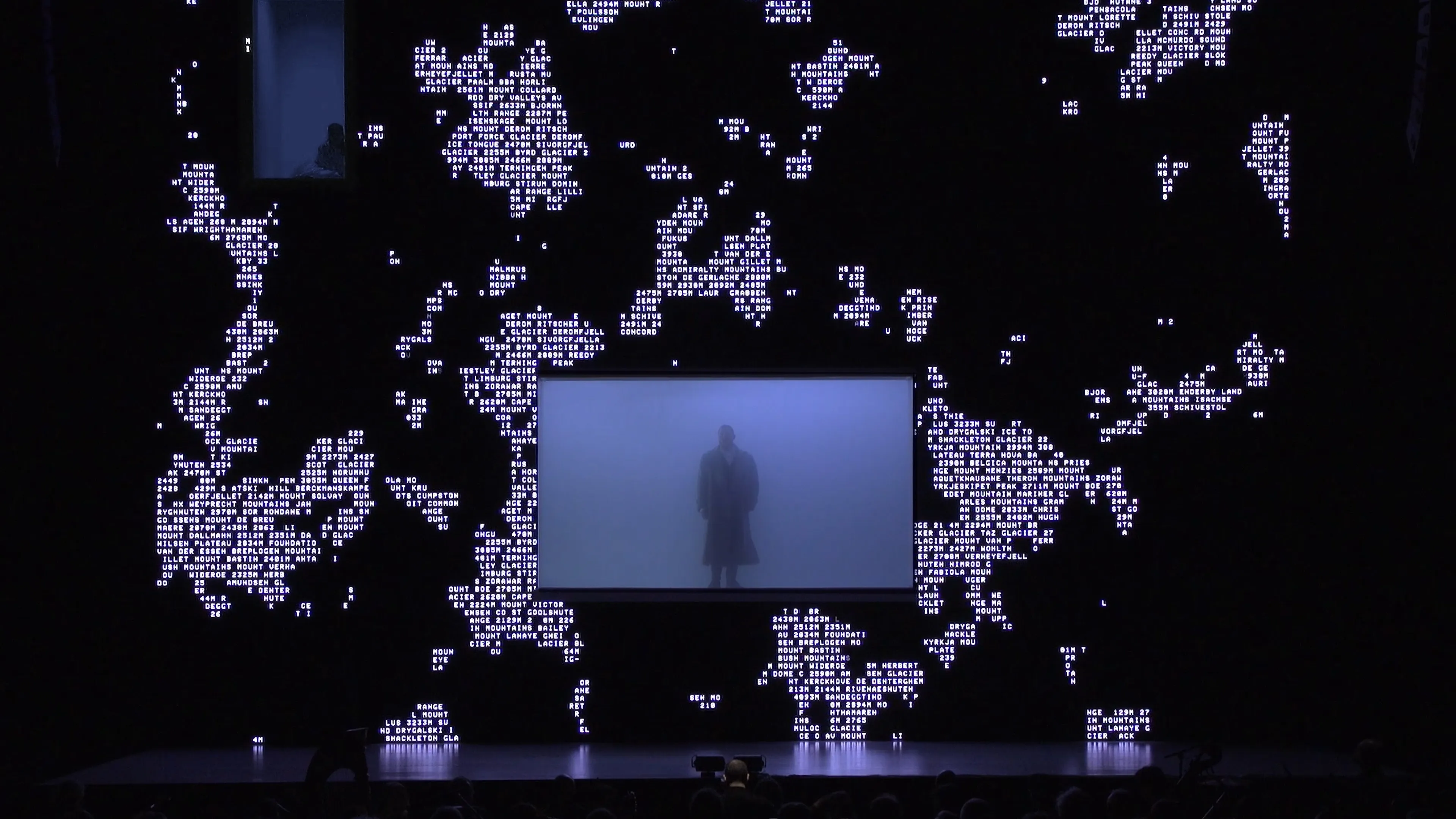 Man singing in a glass enclosure filled with fog and surrounded by a large LED screen displaying white text on black that resembles a map of continents.