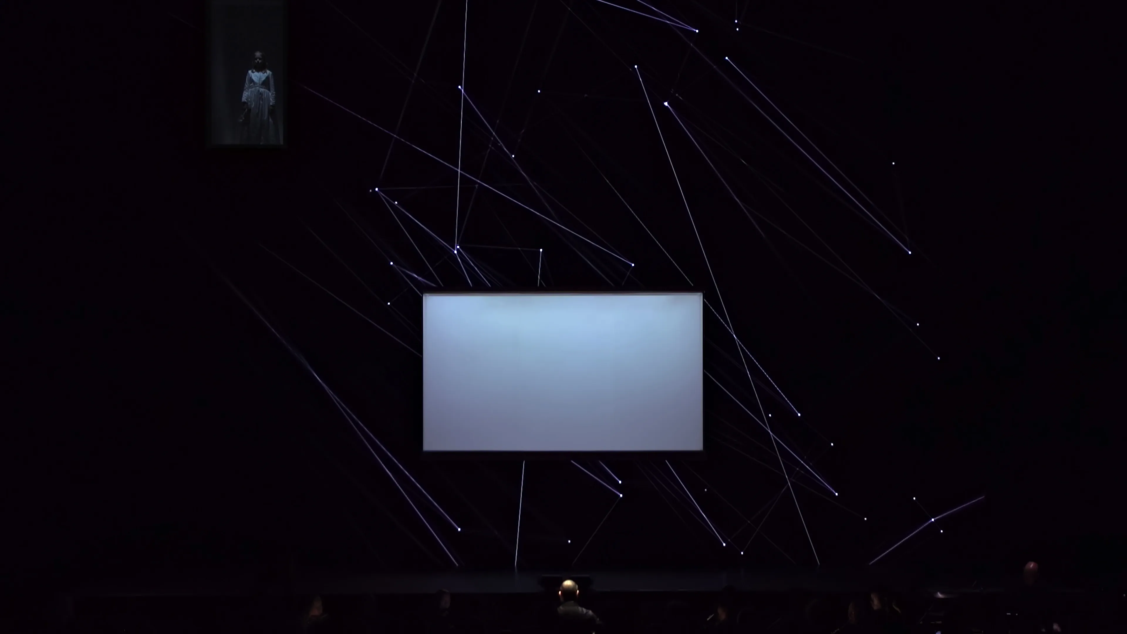A large LED screen on a stage behind an orchestra displaying geometric lines and points. In a cut-out of the screen, a young girl wearing white is visible in top-left.