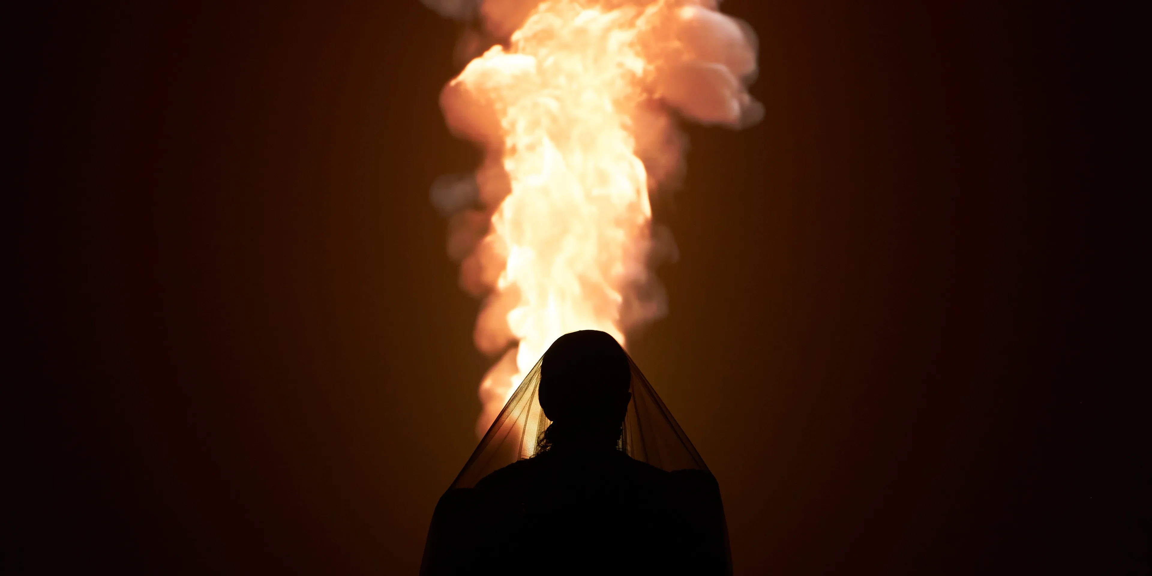 A silhouetted woman wearing a black veil stands in front of a projection of fire.