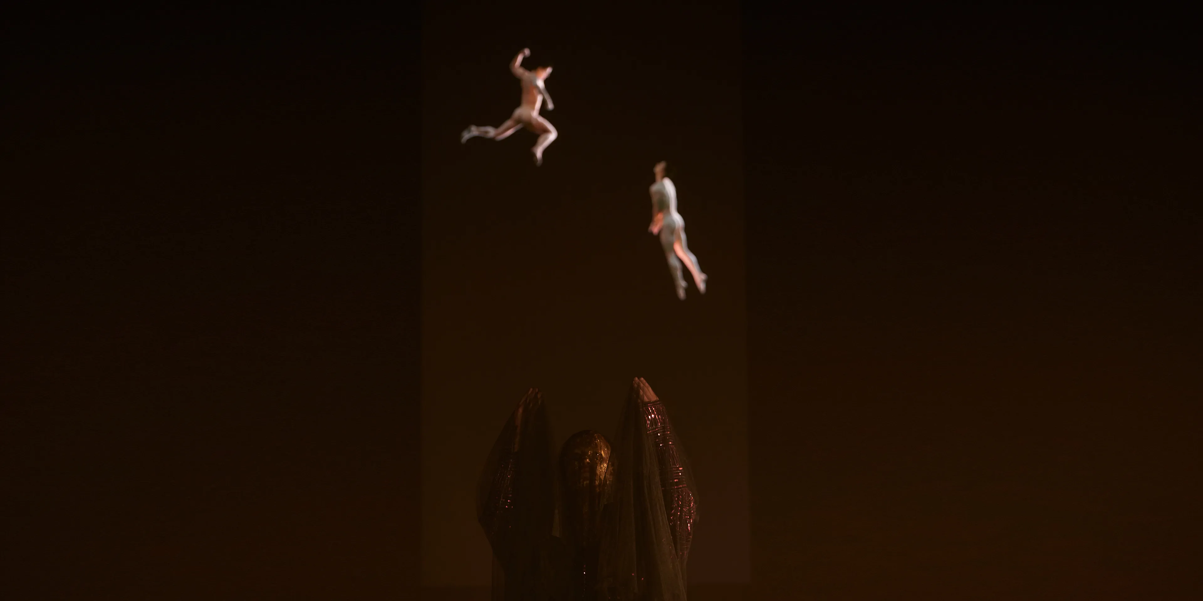 A person wearing a gold mask and a black veil stands with her hands raised in front of a large projection of two male bodies floating in space.