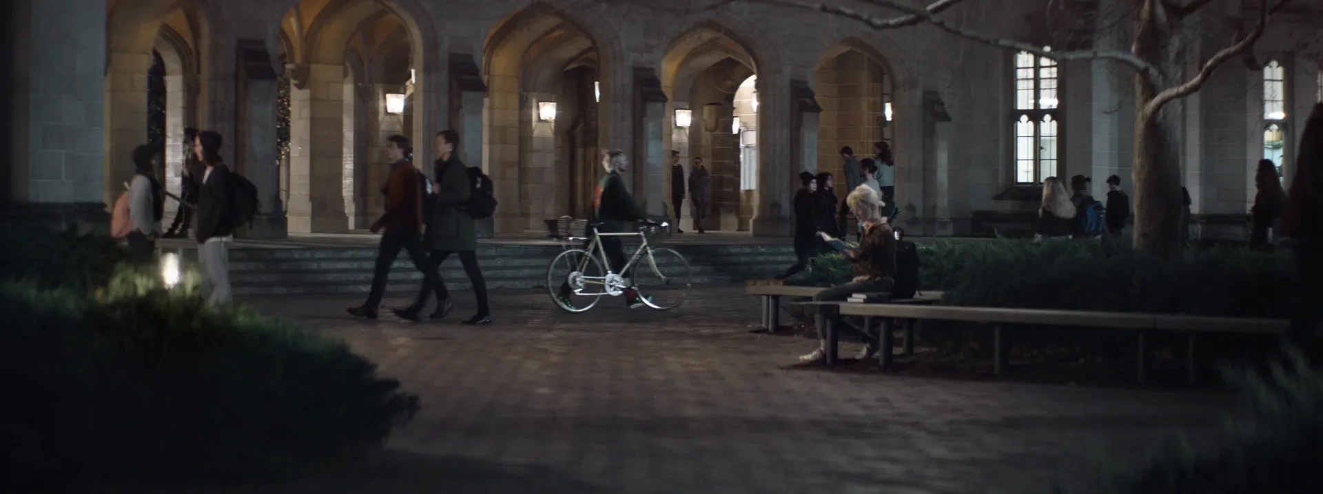 At night, a girl reads a book on a bench in a university courtyard as students walk past.