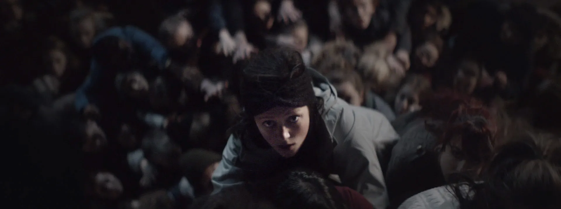A girl climbs a trunk-like structure of bodies. She is looking up above past the camera.