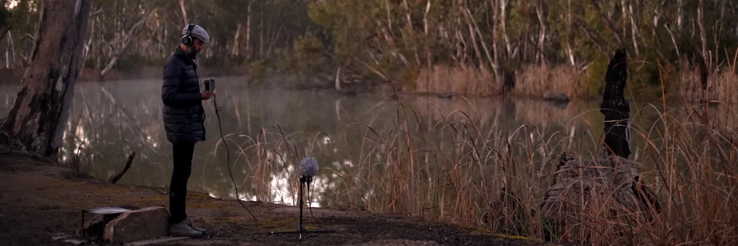 Beside a river, a man wearing headphones holds a sound recording device in front of a microphone on a stand.