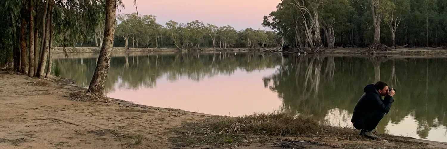 A man is crouching beside a river at dawn, taking a photograph.
