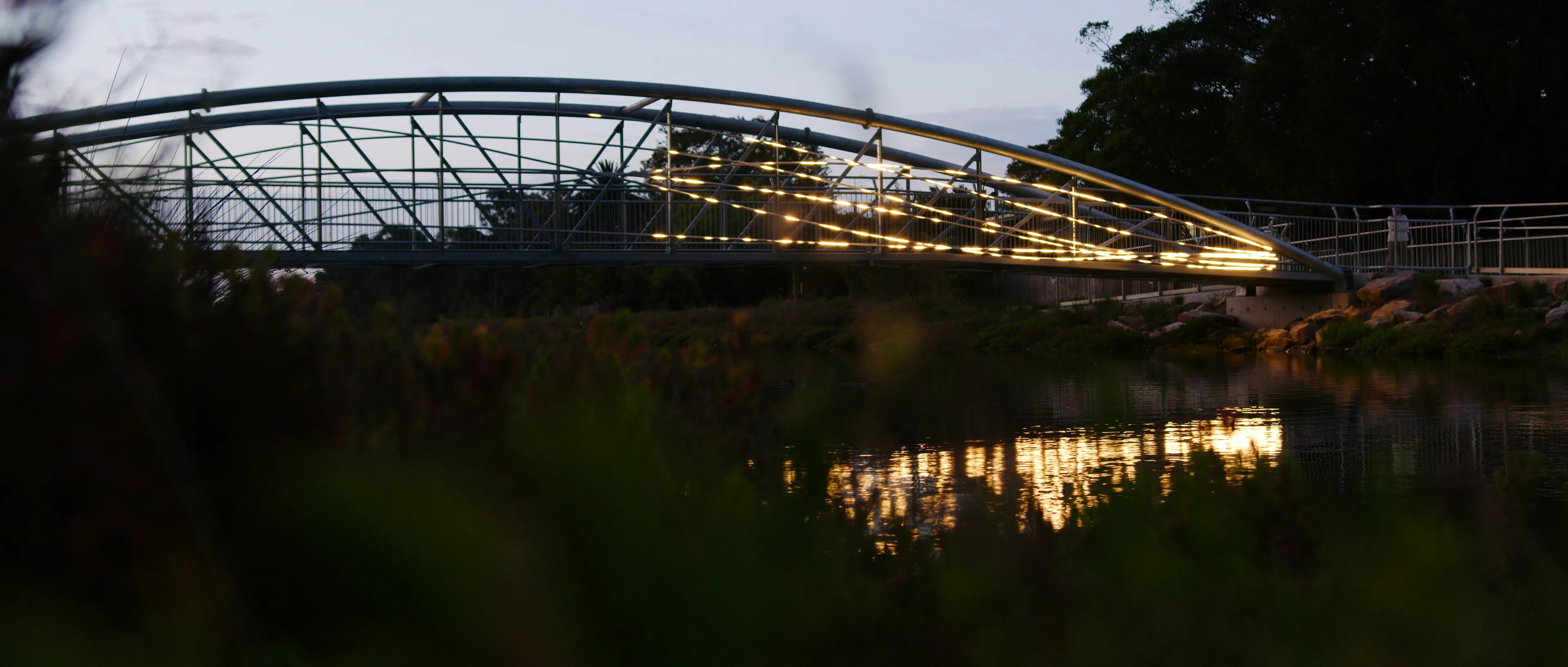 Flyway light installation illuminating a pedestrian bridge over a river at dusk.