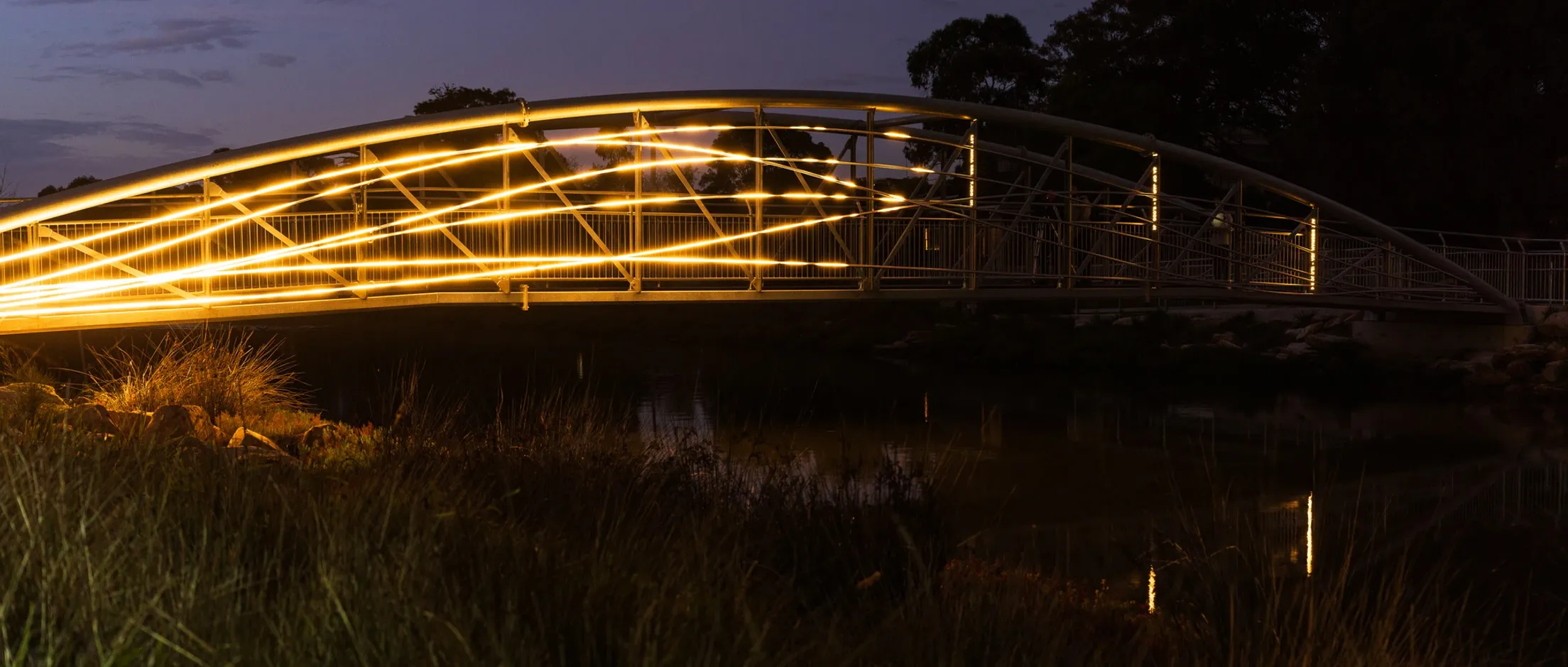 Flyway illuminating a pedestrian bridge with intersecting bands of golden light reflected in the water below.