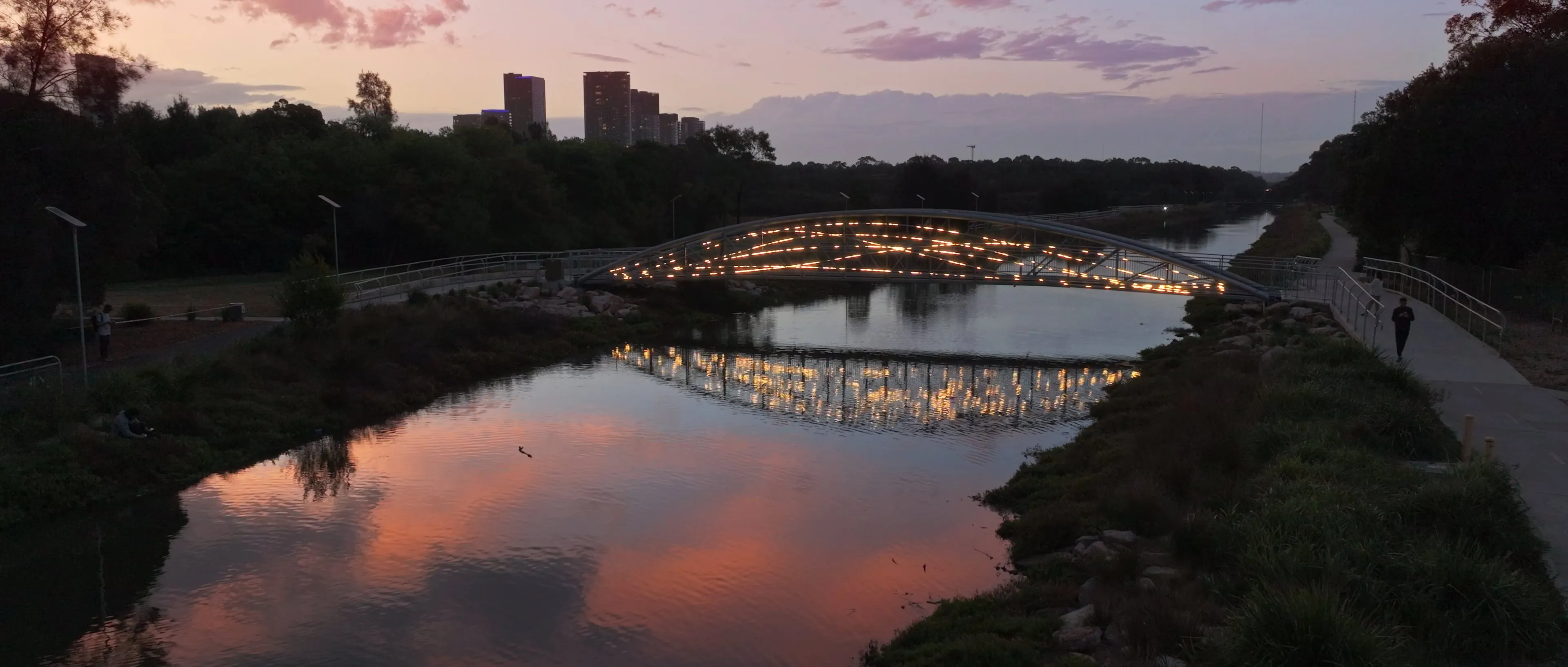 Illuminated light artwork on a pedestrian bridge spanning a river at dusk