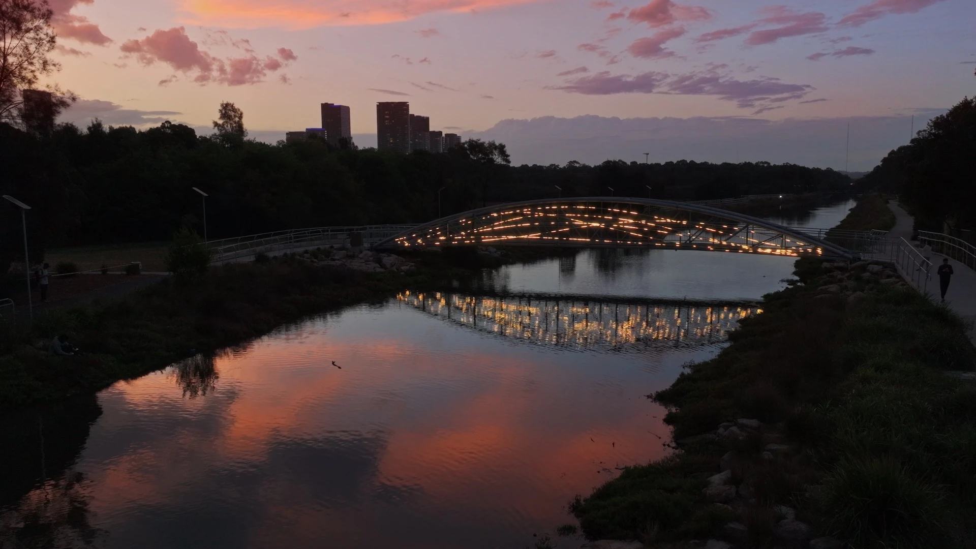 Illuminated pedestrian bridge spanning a river at sunset, with glowing lights reflected in the water.