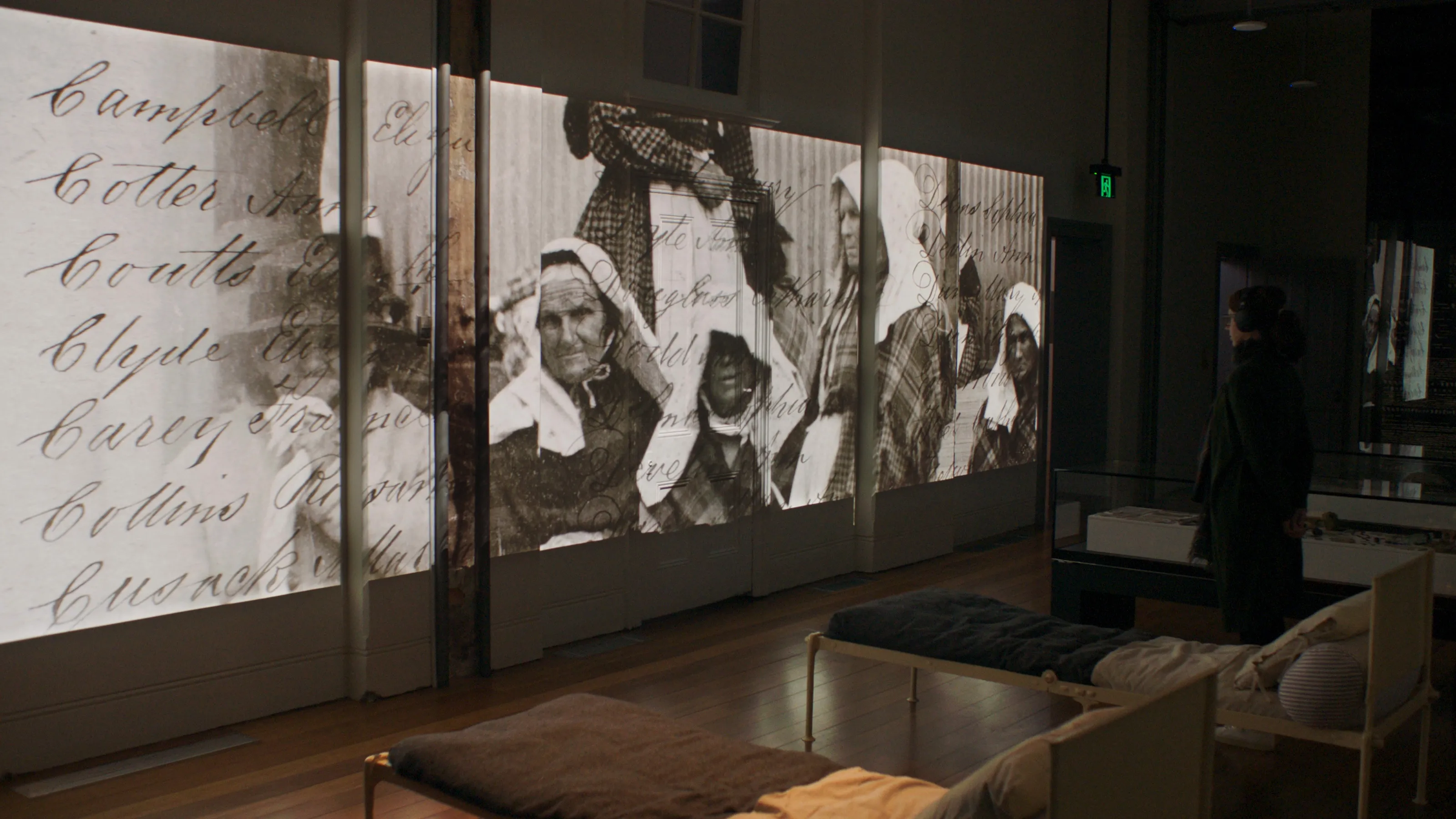 A woman stands in a museum in front of a wide projected image of historical photography of elderly women and handwritten typography.
