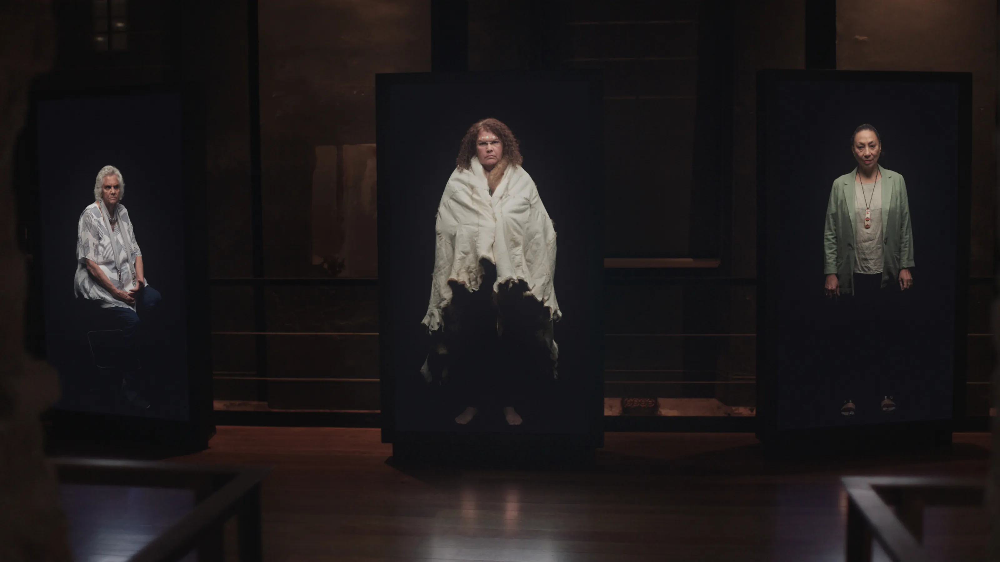 Three large vertical video screens sit side by side in a dimply lit room. Each screen depicts a person at 1 to 1 scale, standing in a black void.