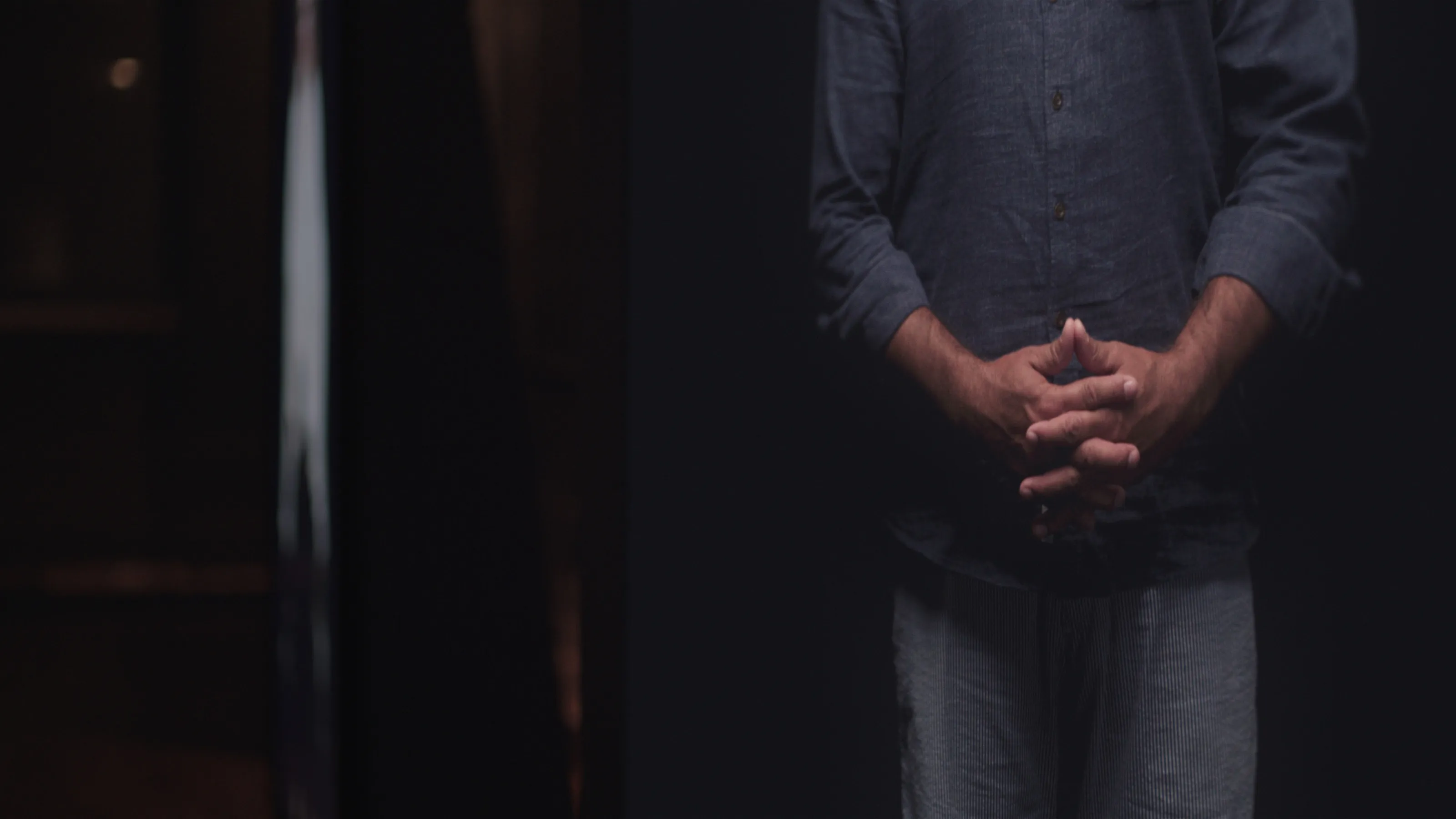 On a video monitor inside a dimly lit room, a man holds his hands in front of him.