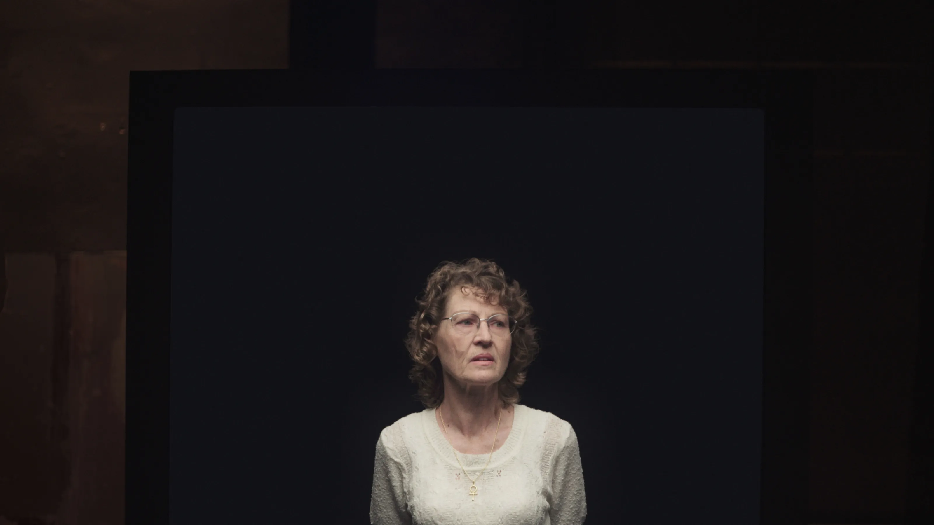 The image of a woman standing in a black void with a concerned expression on her face is depicted on a large video screen in a dim room.