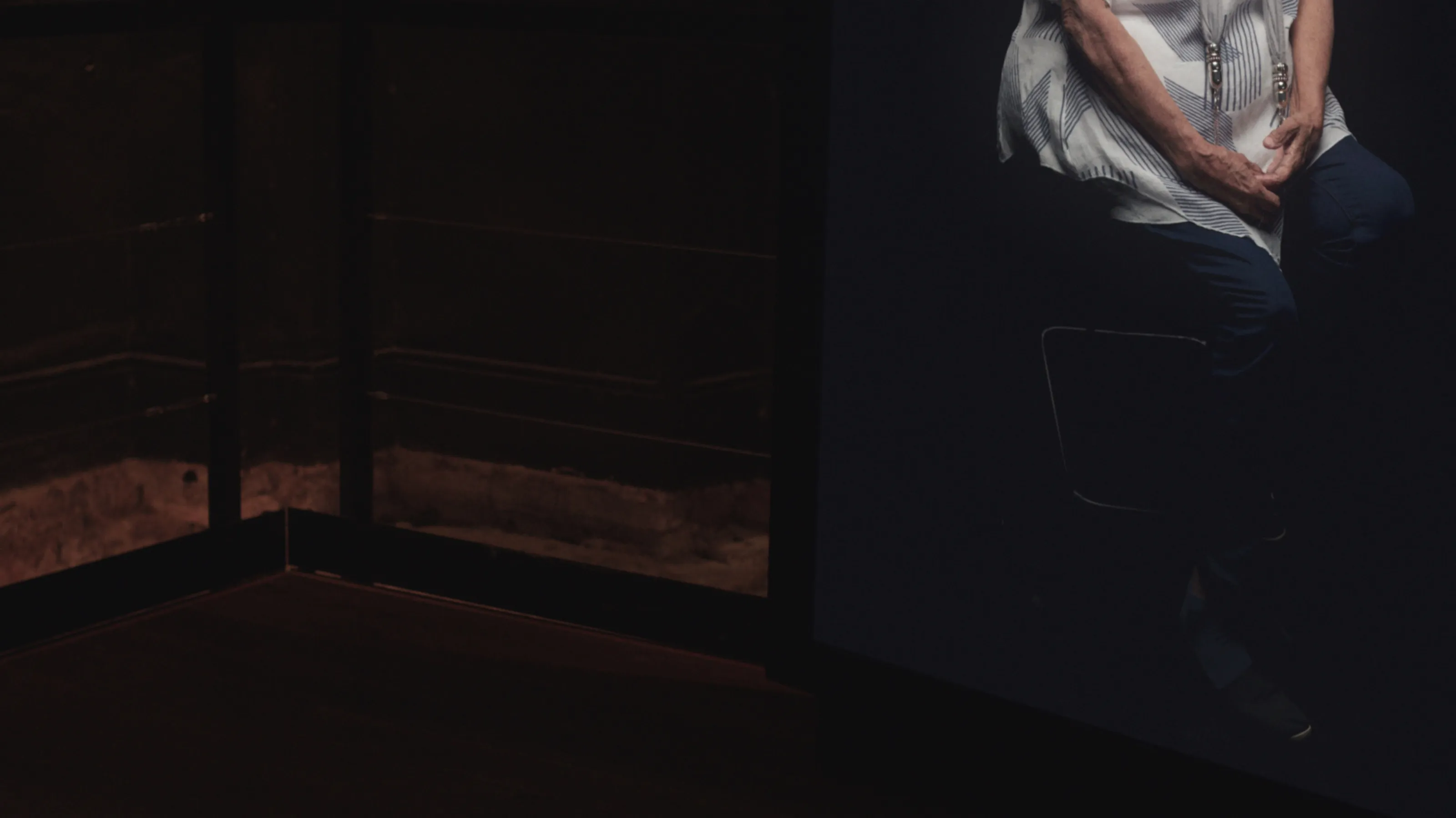Close-up of a video screen in a dim gallery space. The legs of a woman sitting on a chair in a black void can be see on the screen.