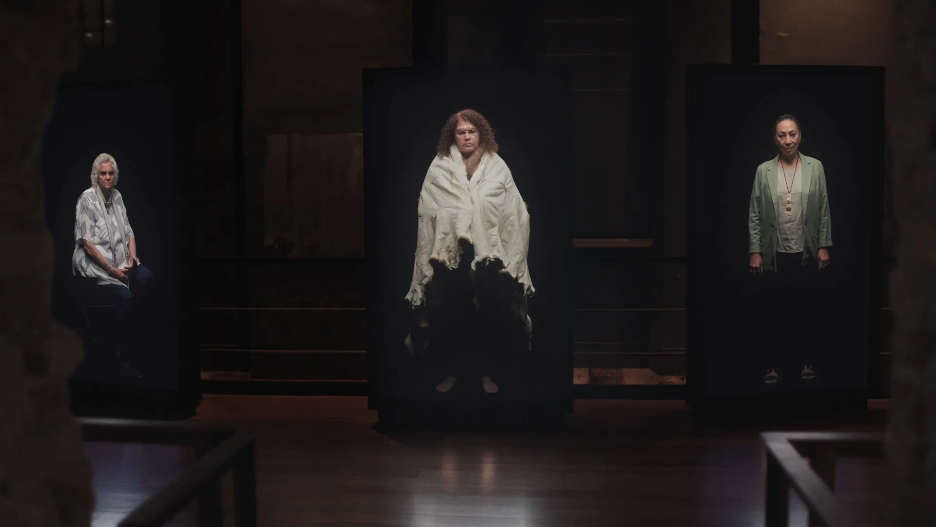 Three women appearing on large vertical screens in a dark exhibition space.