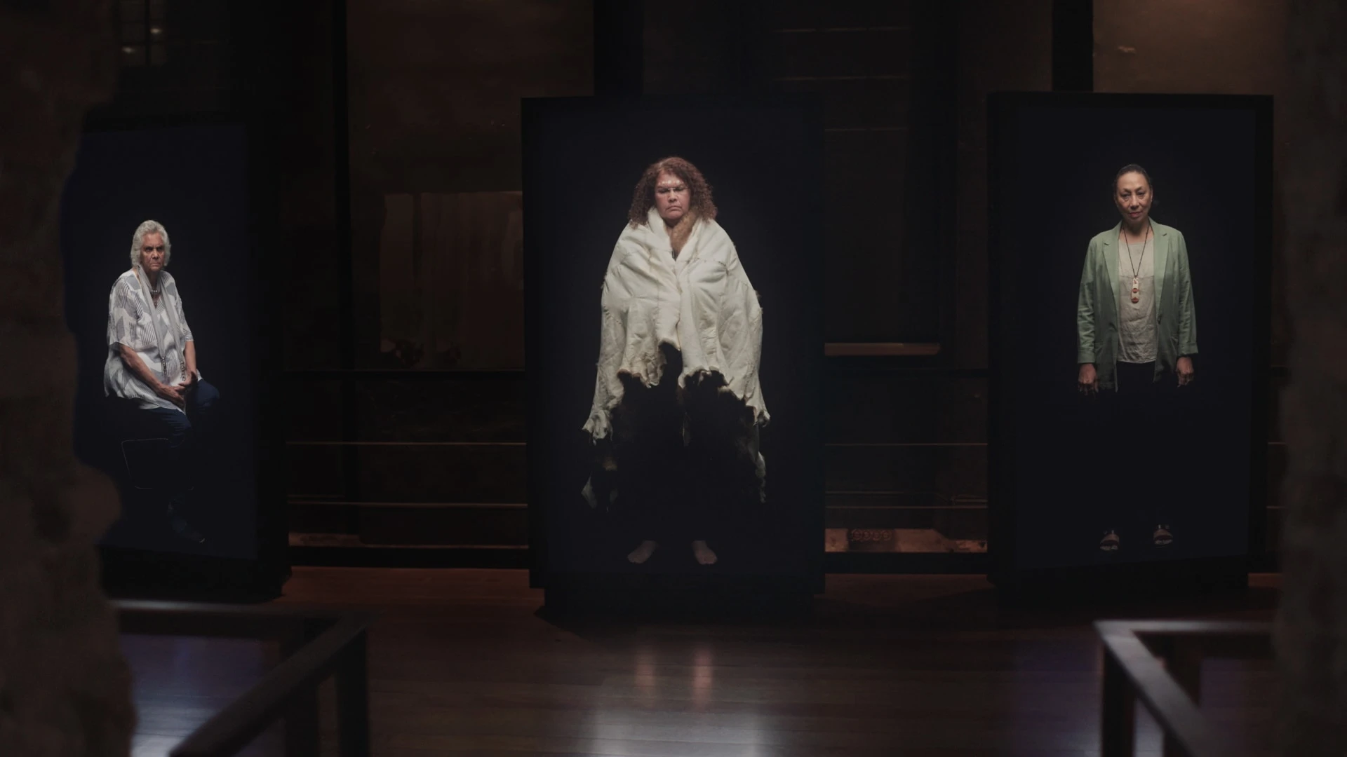 Three women appearing on large vertical screens in a dark exhibition space.