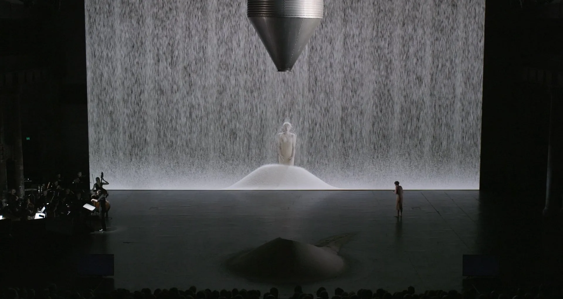 Opera stage with a figure surrounded by falling white particles on a large screen