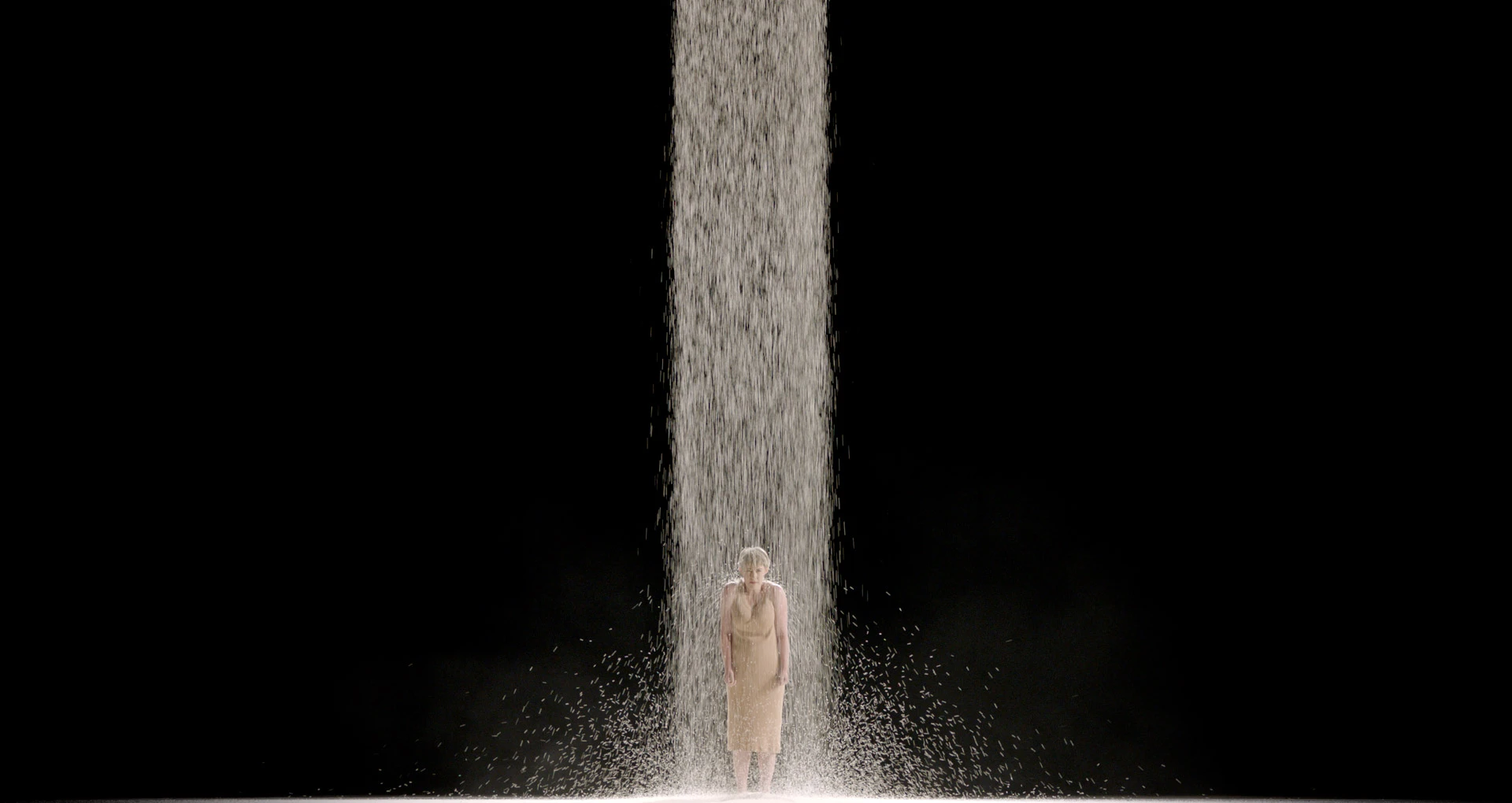 Lone performer standing beneath a column of falling material on a dark stage.