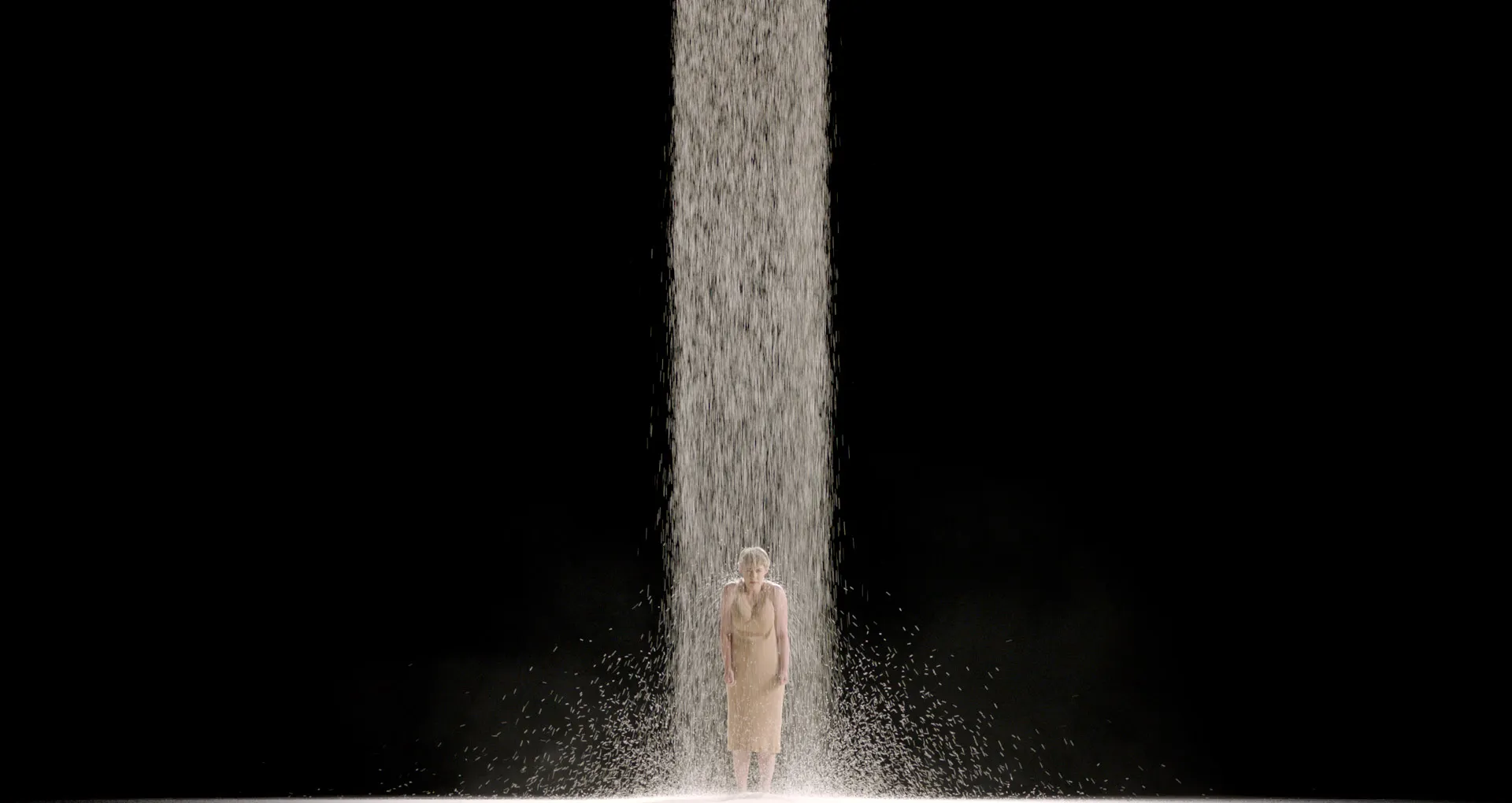 Lone performer standing beneath a column of falling material on a dark stage.