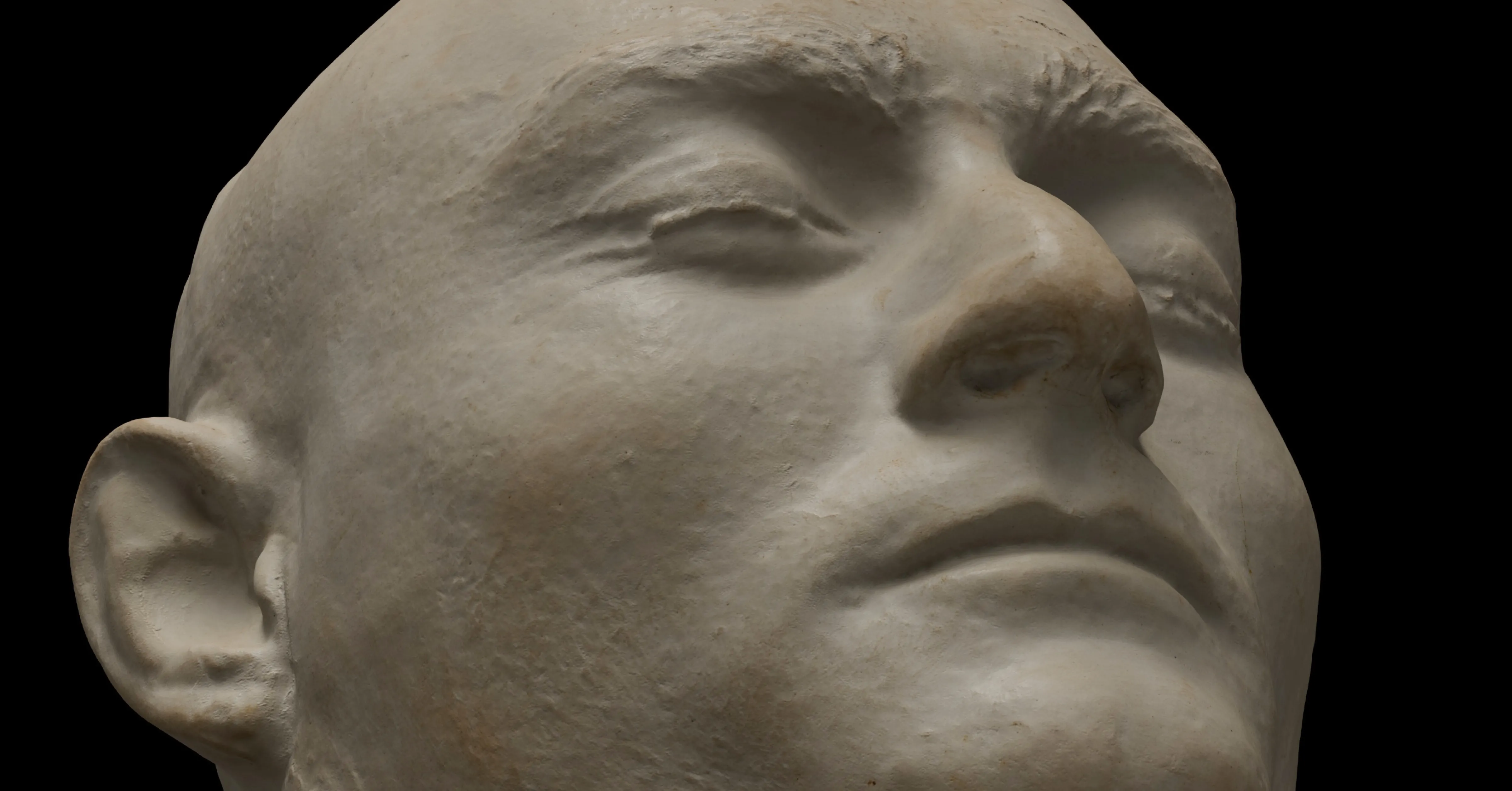 Close-up of a death mask showing a serene face with closed eyes and pronounced facial features against a black background.