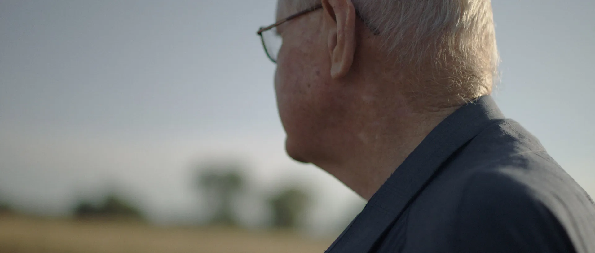 An elderly man stands outside during the day. In the backgreound is an out of focus horizon and some trees.