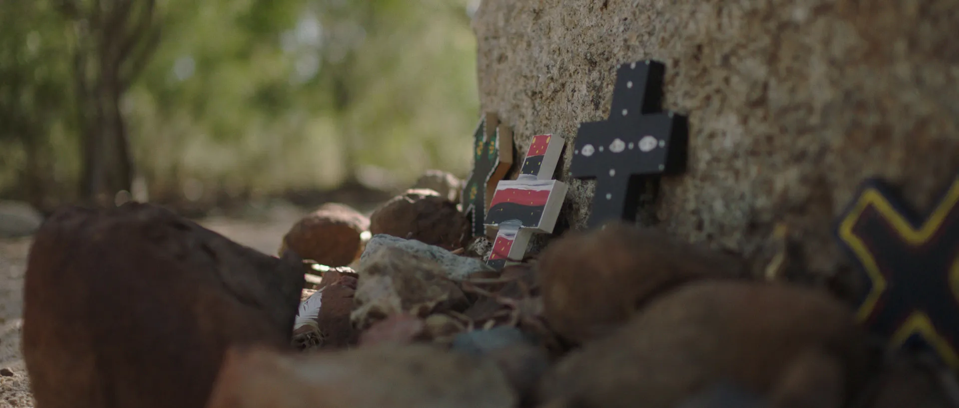 Hand-made, painted wooden crosses sit at the base of a large rock, surrounded by smaller rocks.
