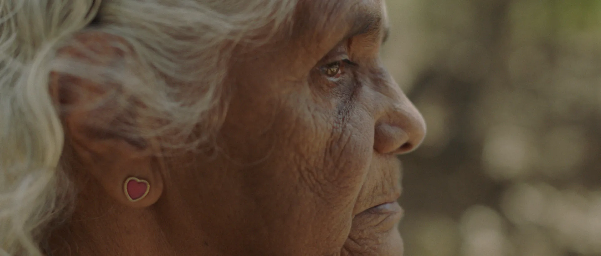 Close-up profile of an older woman with white hair, wearing a small red heart-shaped earring.