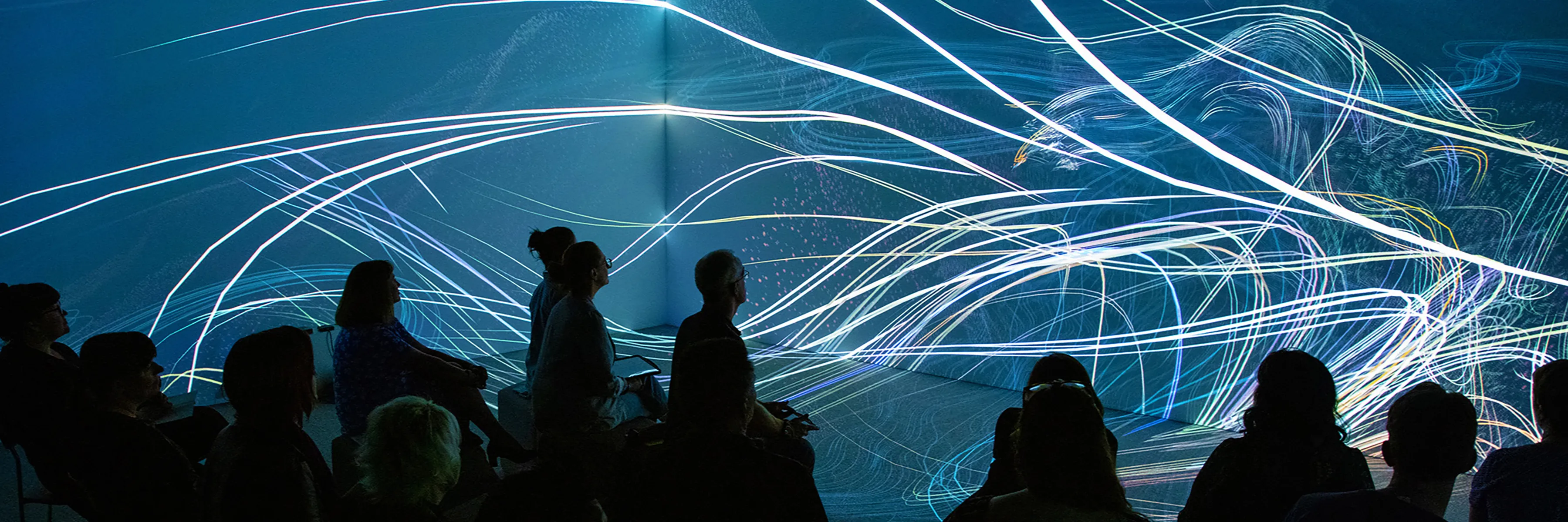 15 people seated in an immersive projection environment watching imagery of white flowing lines and points on a blue background.