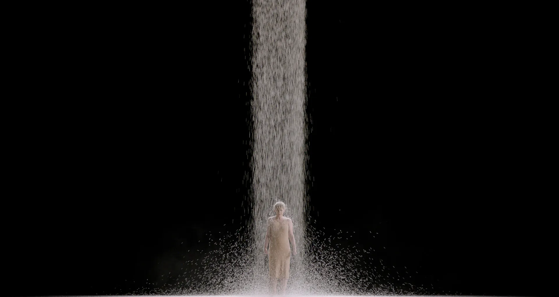 A stream of rice falls on a woman standing in a black void. The rice is piling up around her.