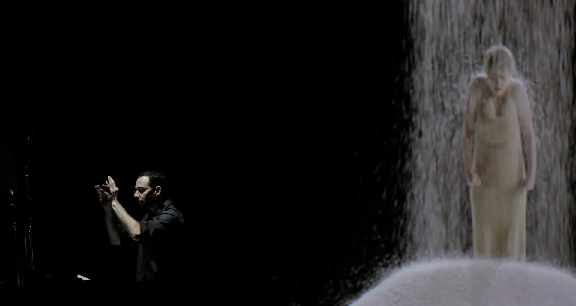 A conductor performs on stage in from of a projected image of rice falling onto a woman.