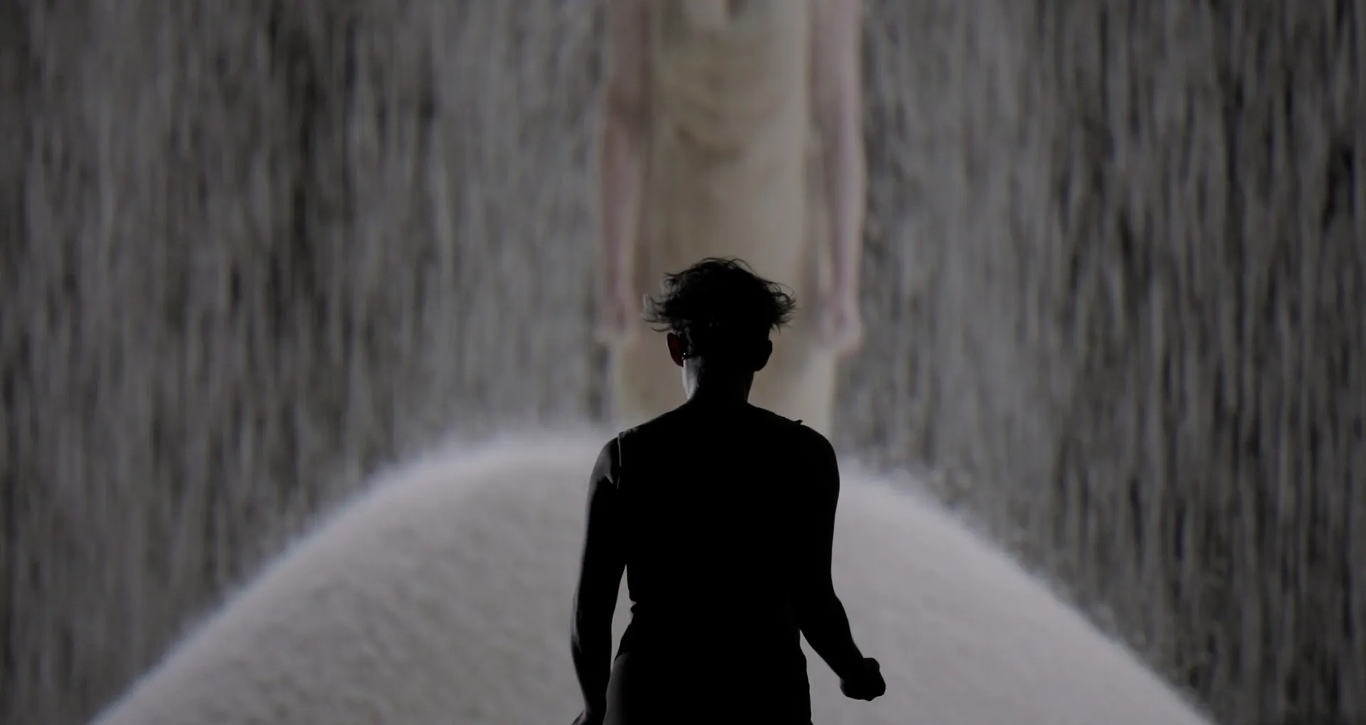 The silhouette of a female singer standing in front of a projected video image of her standing in a pile of rice as more rice falls on top of her.