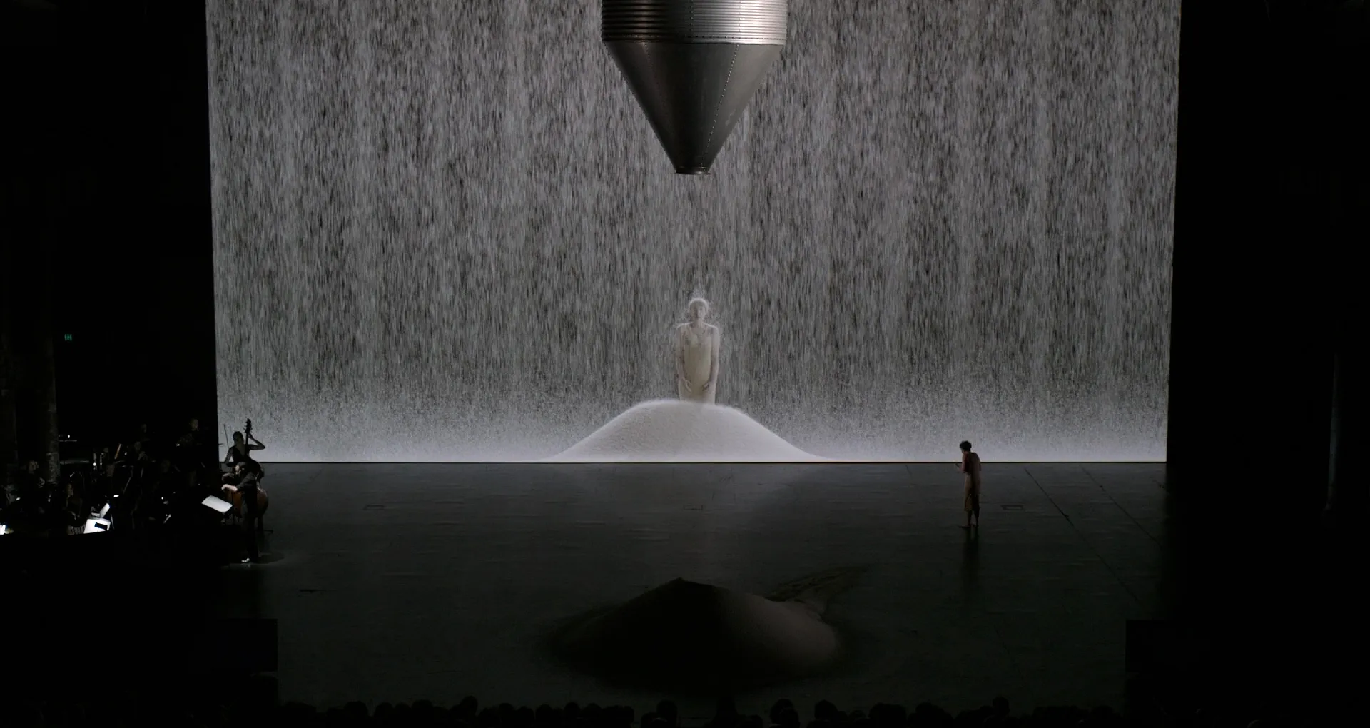 A stage with a performer, an orchestra, a large pile of rice and very large projection screen, which depicts an image of rice falling on a woman.