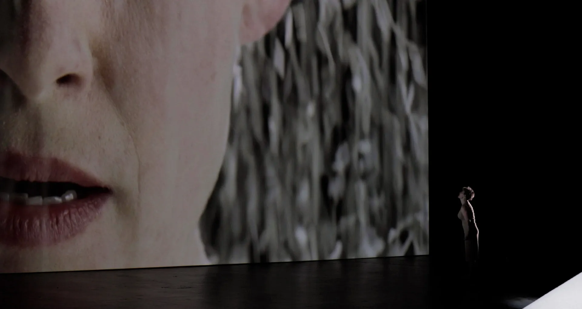 A woman stands on stage in front of a massive projection screen, displaying a close-up image of the same woman with rice falling onto her.