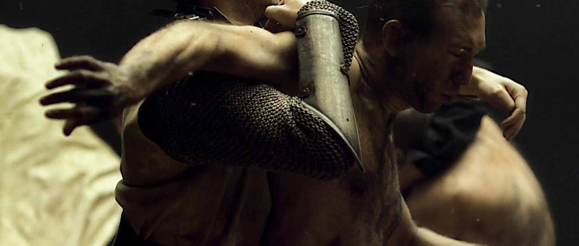 Dirt-covered performers intertwined in a close physical embrace, wearing chainmail and armour