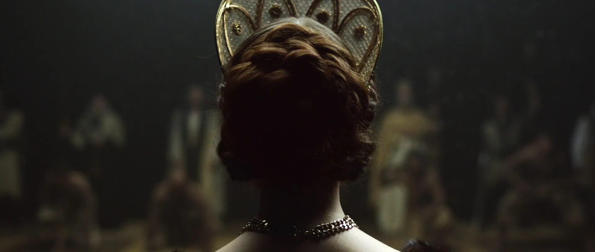 Rear view of a woman wearing an ornate crown in a dimly lit historical scene.