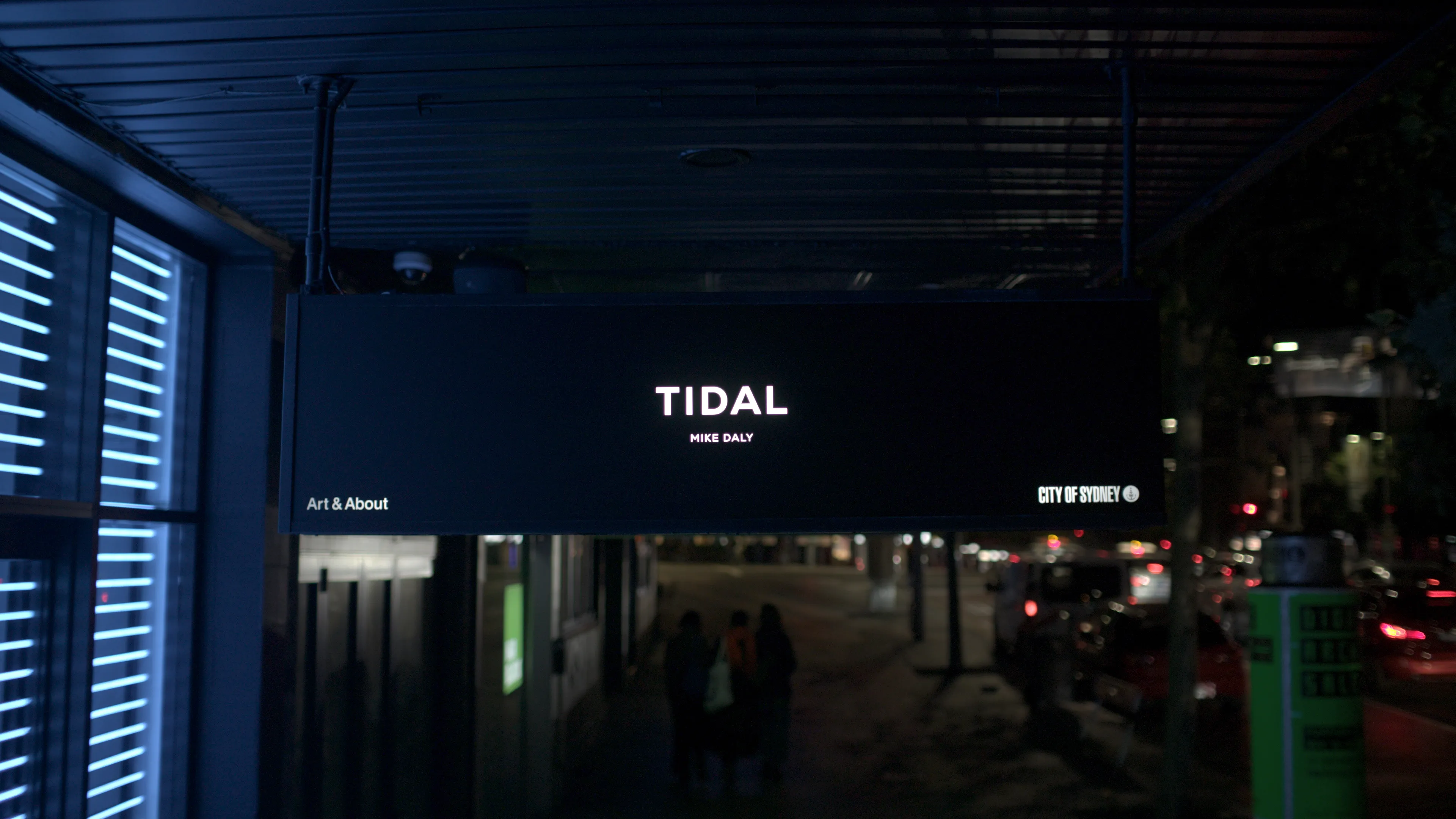 A black under-awning sign on a street at night in front of the light installation artwork. Text on the sign reads: TIDAL, Mike Daly, Art & About, City of Sydney.