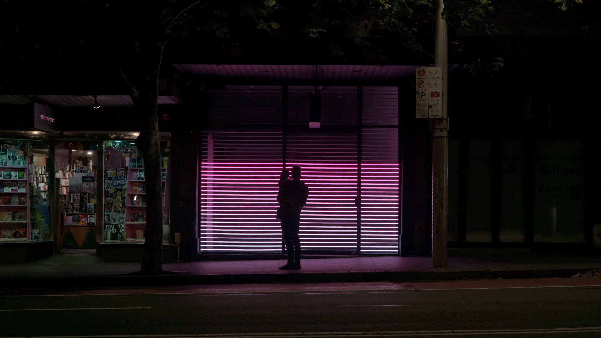 Silhouetted figure holding a child in front of a pink light installation on a city street at night.