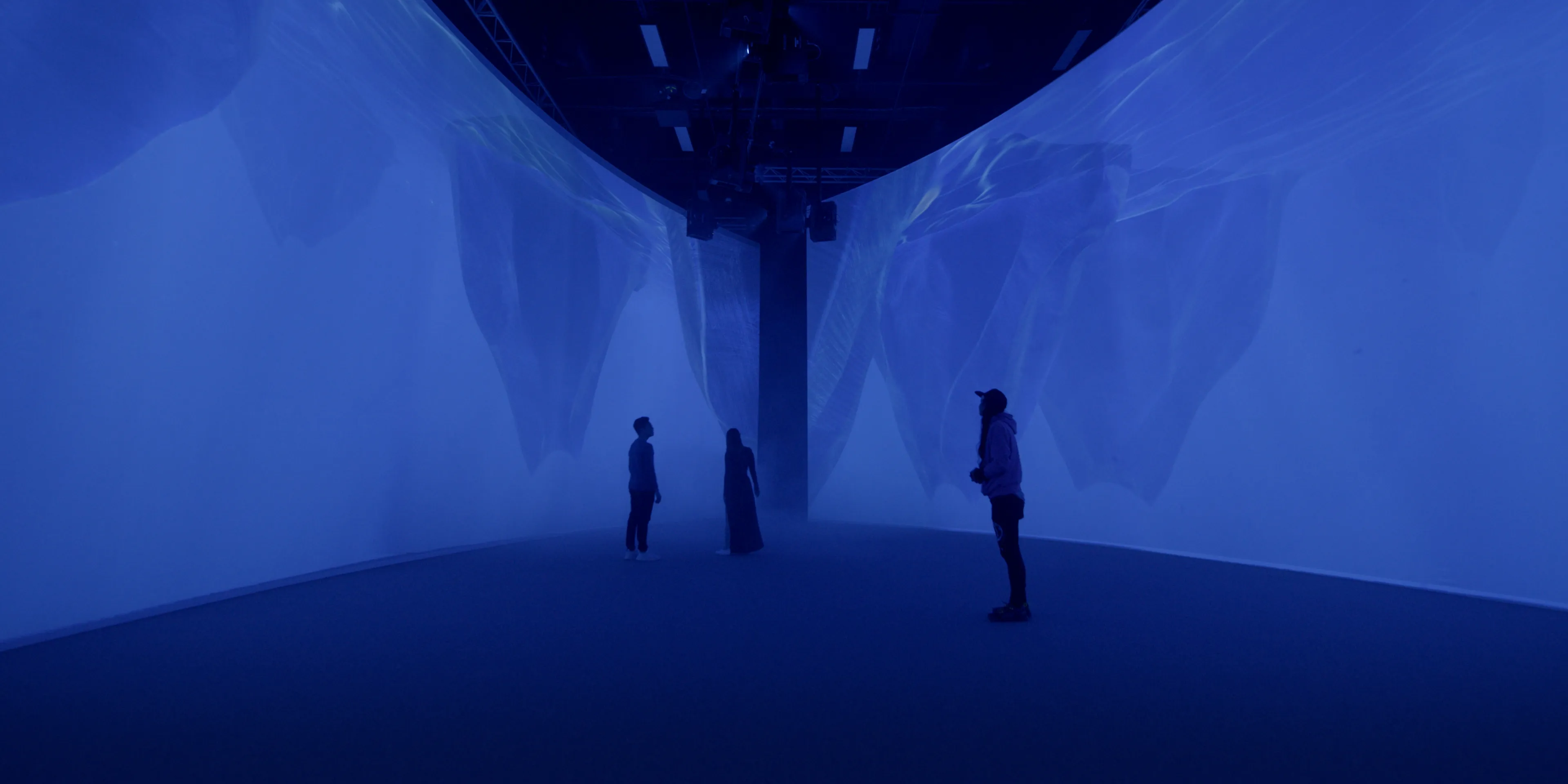 Three people stand in a long immersive projection space with curved walls. the imagery on the walls depicts an underwater scene featuring several large icebergs.