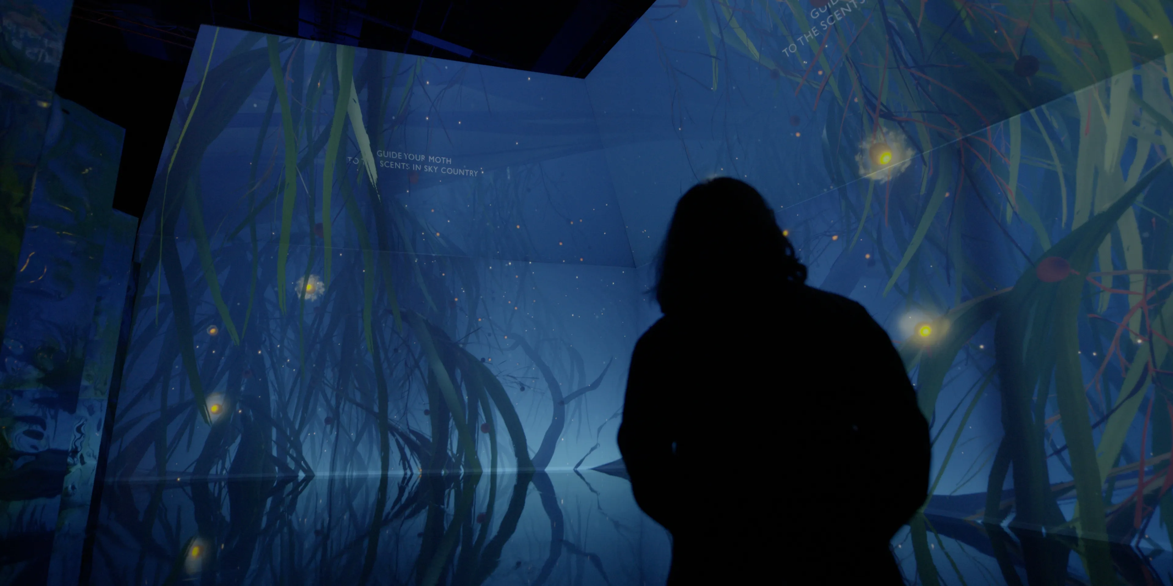 A silhouetted person in an immersive projection space of hanging leaves in a foggy blue void. Text on the walls read 'Guide your moth to scents in the sky country'.