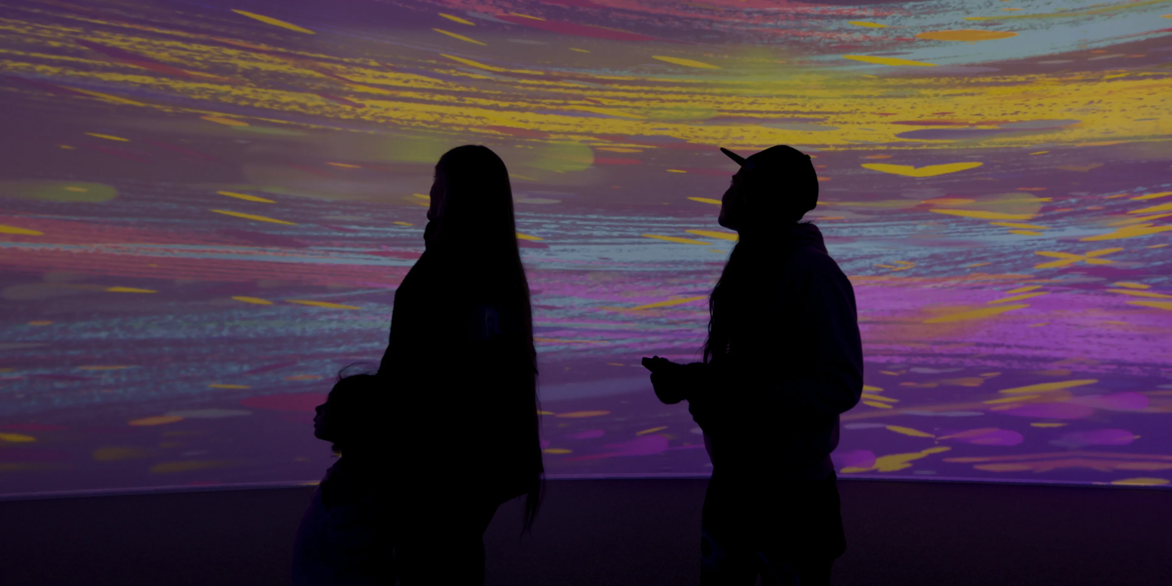 Two silhouetted women stand in front of a large projected image of colourful paint-like streaks.