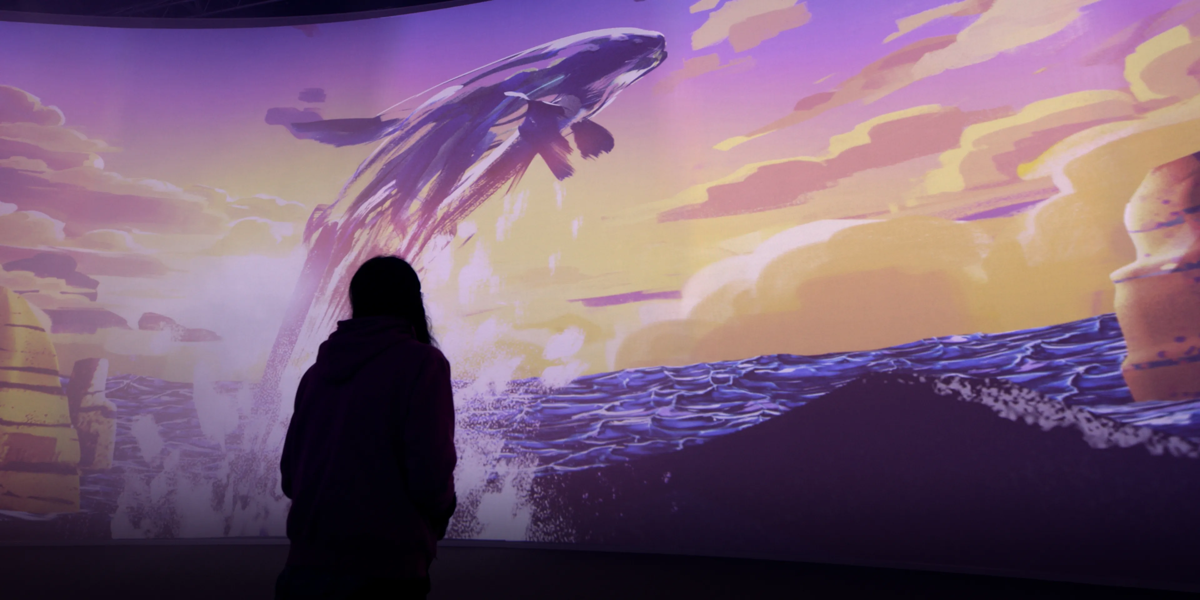 A silhouetted person stands in front of a painterly-looking projected image of a whale launching out of the ocean at sunset.